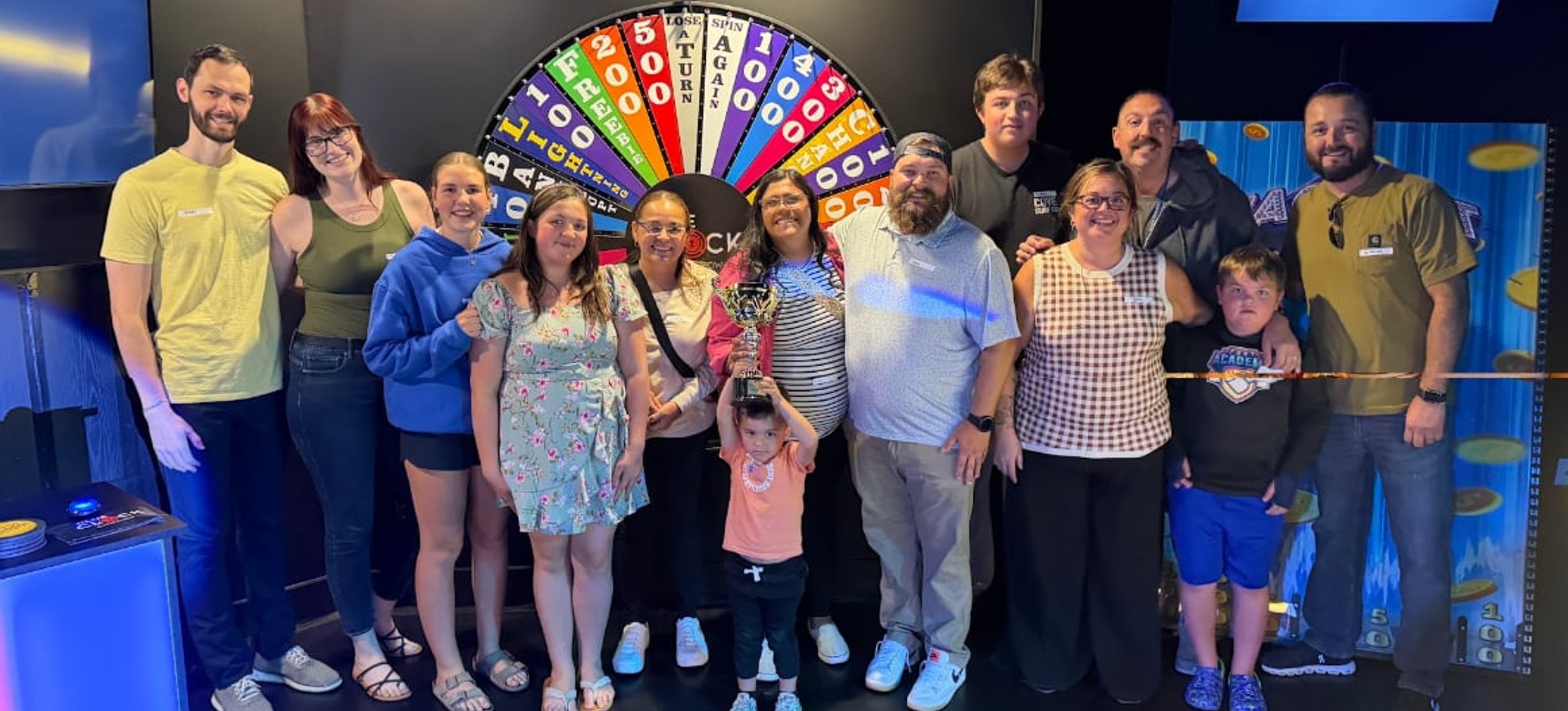 Group posing in front of the Spin To Win prize wheel at ClueClock Gameshow